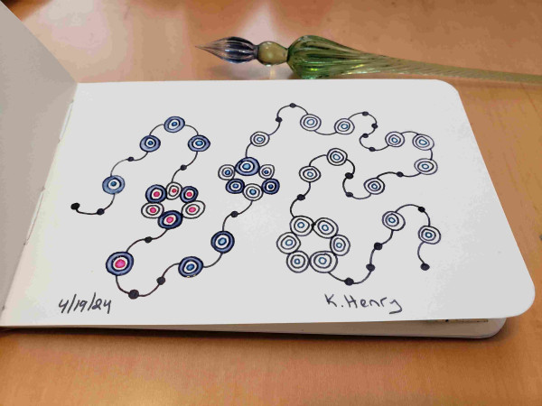 Hand drawn generative art in ink on an open page of my sketchbook. The abstract pattern looks a bit like beads and knots in a string. My glass dipping pen is next to my sketchbook.