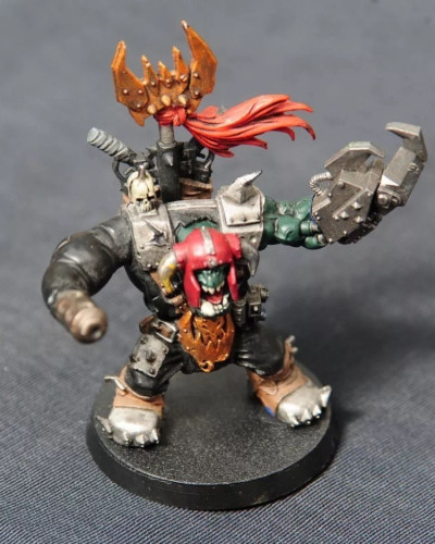 A painted Games Workshop miniature figure of a green-skinned ork in armour, holding a mechanical claw and wearing a red helmet with a red cloth banner.