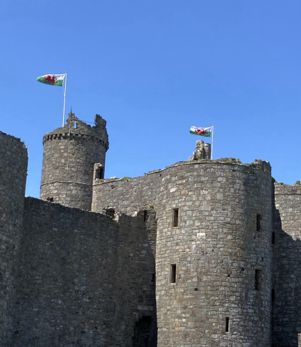 The massive grey walls of the mediaeval castle rear up into a bright blue cloudless sky. From two of the towers the flag of Wales is bright red, white and green.