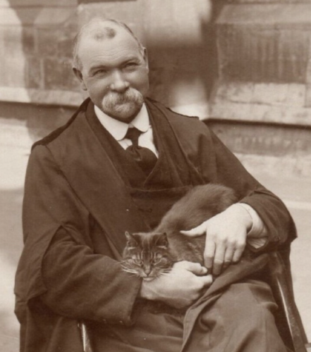 Black and white photo of a smiling, kindly-looking older white man wearing a suit with some kind of churchy black robe garment over it. He's sitting in a wooden chair and cradling a dark tabby cat in his lap. The cat is looking at the camera with a very serious expression.