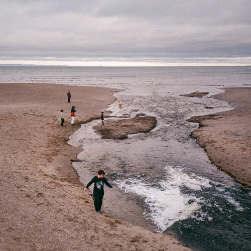 Colour photo of children playing beside a stream flowing strongly across a beach to the Firth of Forth. The sky is greay with a slight tinge of colour, but otherwise the colours are muted.