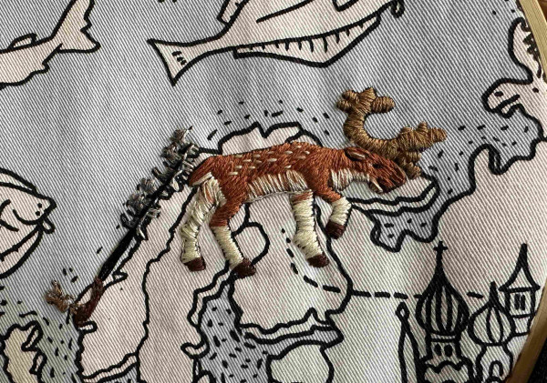 An embroidered fallow dear on a fabric map. Also visible is a team of dogs pulling a sled. 