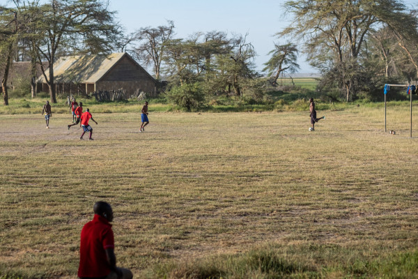 Men playing soccer on a dry grass field with trees and a rustic building in the background.