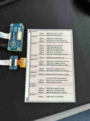 A close up pic of an epaper screen showing the schedule of several different teams (76ers, bucks, PWHL teams, avalanche, etc). It’s laying on a black desk mat and has blue computer-looking parts connected to a gold ribbon coming out of the left side. 