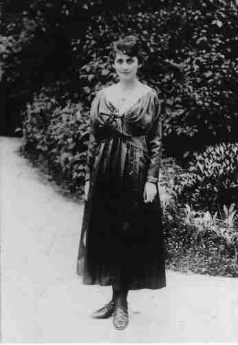 Black and white photograph of a woman photographed in a park or garden. She is standing on a path. Behind her, the leaves of shrubs and trees are visible. The woman is dressed in a refined pleated dress of dark colour. Her hair is dark and neatly styled. The woman is standing in the centre of the frame.