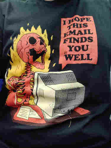 A selfie closeup shot of a black t-shirt with a red human skeleton on fire, sitting at a desk with a computer that says "I hope this email finds you well"