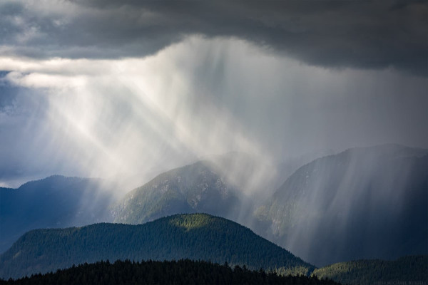 A storm cloud dumps rain on the slopes of a few mountain peaks while sunlight breaks through the clouds to the left and illuminates the rain and mountains.