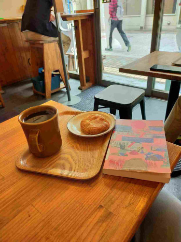 The photo is from inside the cafe where the legs of someone can be seen outside the window. On a brown wooden table is the pink paperback book with illustrations representing people & things around Tokyo Ueno Station such as a panda & train station facades. To the left is a brown wooden tray with a cookie on a white plate & a brown mug of black coffee. In the distance the back of a Japanese man can be seen sitting on a stool at a counter looking out the window