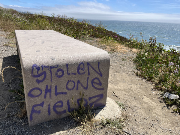 A concrete bench on a hill overlooking the ocean. The side of the bench is spray-painted with the words “Stolen Ohlone Fieldz”.