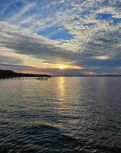 Looking out across a vast river at sunset as the bright yellow sun's orb lowers at the horizon, casting shades of yellow and orange across a blue sky nearly blanketed with white cloud formations. The sun beams a column of light across the water's surface as it vanishes behind the horizon's low clouds.