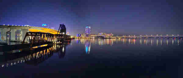 Panoramic view across a calm, dark river, where a large concrete bridge and small train trestle cross the water's, illuminated by colorful lights. The opposite shoreline with a city skyline and a variety of colors, all reflecting upon the water below.