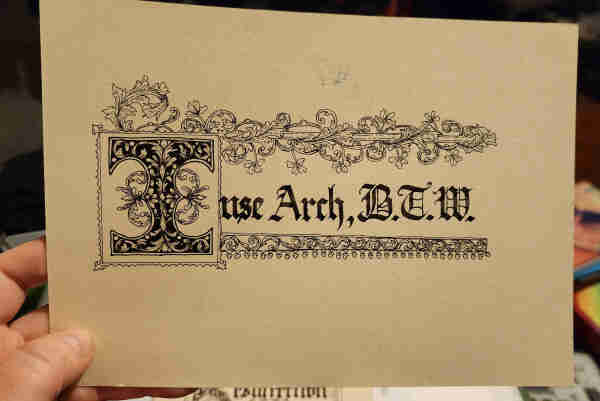 An A5 piece of cream paper with 'I use Arch, B.T.W.' calligraphied on it in blackletter script blank ink. The initial 'I' is big and decorative, and the calligraphy has a scrollwork border across the top and bottom, with swirling leaves and vines.