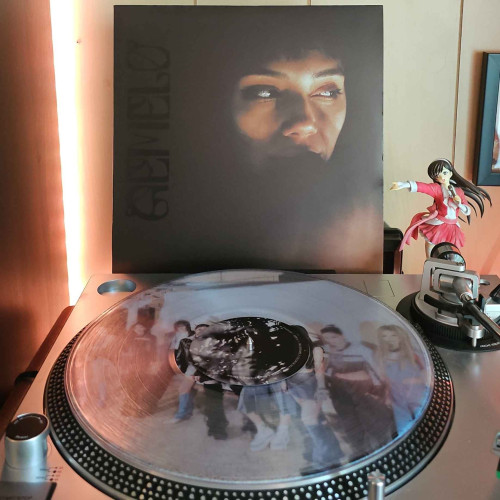 A clear vinyl record sits on a turntable. Behind the turntable, a vinyl album outer sleeve is displayed. The front cover partially shows Angelica Garcia's face as it fades into a dark background. 