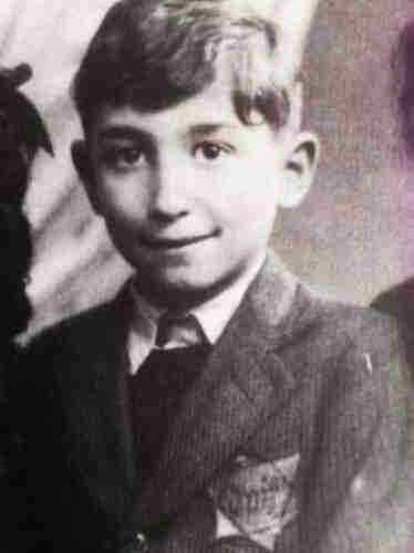 The boy in the jacket. He has a six-pointed star sewn onto his chest with the inscription Juif. He has medium-length hair and a fringe falling over his forehead. His eyes are wide open and he smiles slightly. 