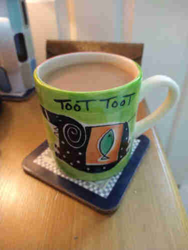 Mug of tea on a bedside cabinet, a white handle to the right, the mug has a green background the words Toot Toot in black text at the top, there's a strange looking black elephant in the centre wearing an orange saddle with a green fish on it 