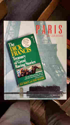 A photo of a paperback resting on a much larger coffee-table book, as follows:

The large book is...
PARIS – past and present by M. ERCOLE POZZOLI.
The cover photo features a raised concrete planter in urban Paris on a rainy day. The ground is wet and the iconic legs of the Eiffel tower are hazily visible in the background.

Laying on the large book is a green soft-cover book.
The DICK FRANCIS Treasury of Great Racing Stories.
Classic tales of horse-racing by fourteen master storytellers.
Edited and Introduced by DICK FRANCIS and JOHN WELCOME.
"These are rollicking good stories, sure to stir anyone with a drop of sporting blood." - Booklist.
FAWCETT CREST.
A photo near the bottom features a motion-blurred image of jockeys on horseback, racing for the finish line.