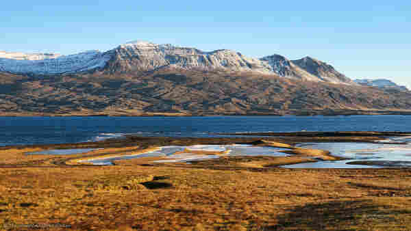 A photo of a landscape under a clear sky and lit from the right. The foreground is brown vegetation with scattered pools of water which have frozen over and are rimed with white ice. In the centre of the shot is a fjord, reflecting the blue sky. On the opposite bank is a chain of mountains, near the centre is one with a prominent peak. The summit is snow-covered but the flanks are brown rock, the texture of the lower slopes is an odd criss-cross pattern. This crosshatch is highlighted by strong shadows. A few white buildings are visible at its foot near the centre left, their size gives scale to the shot.