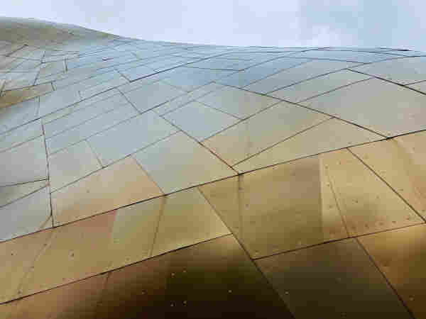 Looking up at a building exterior made of metallic irregular polygonal shapes fitted together over an irregularly curving surface. Closest to us, the colors are bronze and copper tones, and they look more blue as they go toward the sky. 