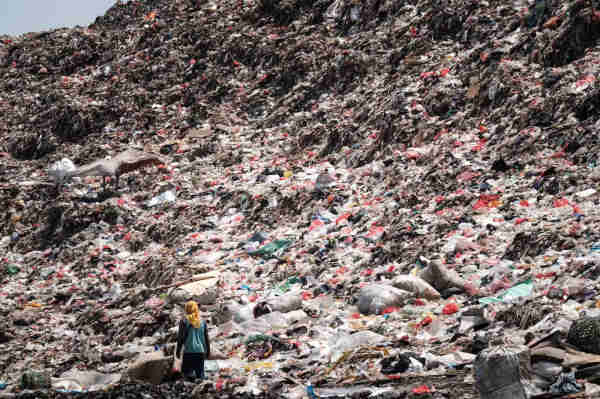 A scavenger, who collects plastic waste to sell, walking in a landfill in Indonesia, a literal mountain of trash mainly composed of single-use plastics.
