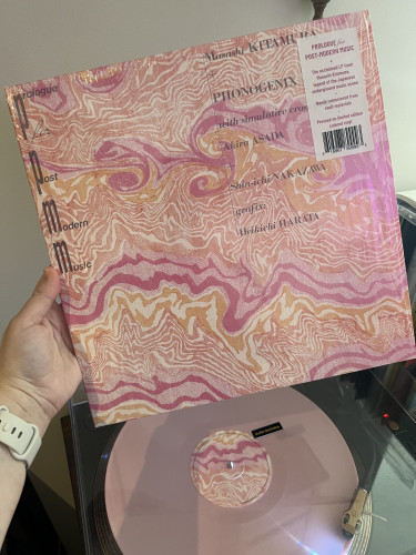 My hand holding up the album sleeve for "Prologue for Post-Modern Music" by Masashi Kitamura and Phonogenix. The pepto-bismol (light pink) coloured record is playing underneath on a turntable 