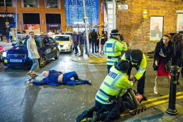 A man lies on the street at night reaching for a bottle of beer as police officers try to subdue another man on the ground. Revelers and other onlookers, along with cars, fill the frame.