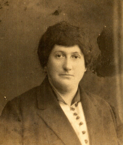 Vintage sepia-toned portrait of a mature womanwith a serious expression, wearing a dark suit with a high-buttoned jacket. The background is nondescript, and the photo shows some age-related damage.
