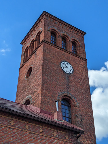 Prostokątna wieża kościoła z czerwonej cegły z zegarem i białej tarczy. 