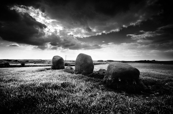 Black and white photo of a row of three standing stones in a grassy field, under a dramatic sky.