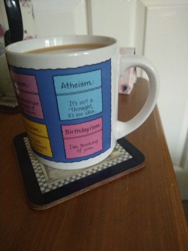 Mug of tea on a bedside cabinet it shows a blue square at the top with a small description of Atheism, below is a pink square describing Birthdayism 
