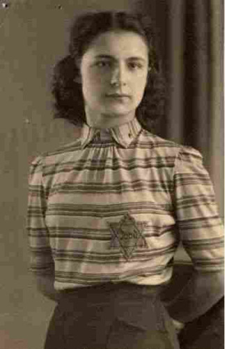 Photo of a teenage girl - from the thighs up. She is wearing a blouse buttoned up to the neck in vertical lines. A six-pointed star with the word "Jood" (Jew) can be seen on her chest. She has long, curly hair pinned back. 