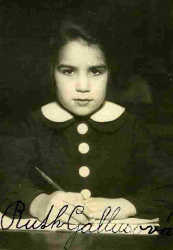 A young girl photographed sitting at a desk. She is holding a pen and holds her hands on some pieces of paper. She is wearing a school uniform wieh large buttons. She has dark hair.