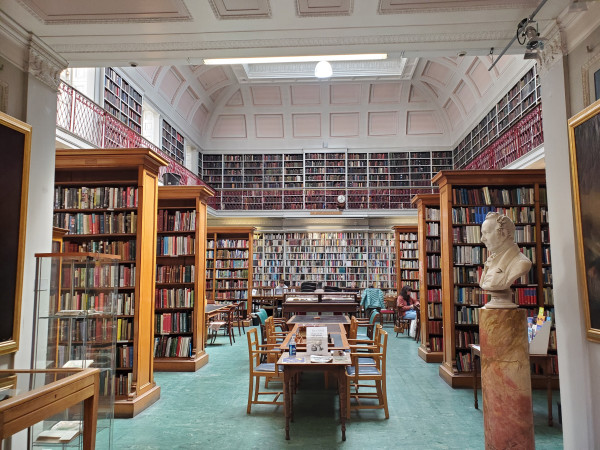 The library of the Newcastle Literary & Philosophical Society. The room has a high ceiling and is well-lit by daylight. On either side 3 large wooden stacks of books. The wall opposite is lined with books, and a mezzanine near the ceiling holds more books. To the left is a marble bust of a Victorian Gentleman on a pedestal. In the centre of the room are long tables and chairs. A few people are sat at them.