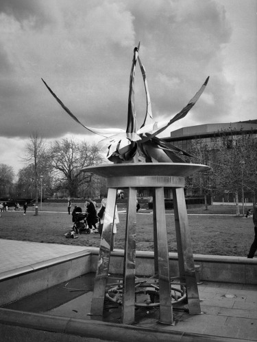Black and white portrait format photo showing a stainless steel sculpture of two swans greeting. They are on a four-legged plinth set in a pool. There is a group of people chatting on the lawn behind. Beyond them to the right is the bulk of the RSC Theatre, under a dark cloudy sky.
