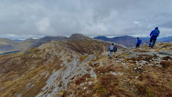 Wide photo of a mountain scene, with four figures walking in front of me along a rocky, heathery ridge between a peak behind us and one in front. The visible ground is mostly in shades of grey, brown, and yellow. In the background are a half-dozen other mountains in the Twelve Bens range in west County Galway, Ireland, with a shelf of grey cloud resting on their summits.