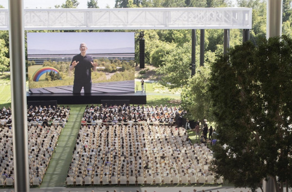 photo of Tim Cook on a gigantic outdoor screen presenting to people at Apple headquarters. There are hundreds of seats looking on - the only thing missing is a woman athlete running up the middle aisle with a hammer. 
