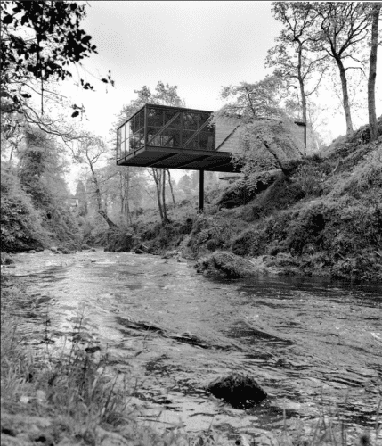 Modern glass-enclosed structure suspended over a small river and embankment. 