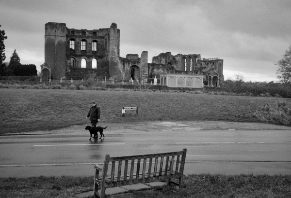 In the foreground, a wooden bench; across the wet road, a man walking his dog, and behind them on the skyline are the stark ruins of Kenilworth Castle. Black and white photo.