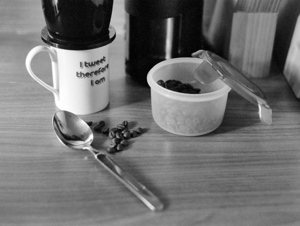 Black and white photo showing the makings of coffee on a bench: spoon, beans, grinder, and black filter/diffuser sitting on a mug, which carries the outdated message "I tweet therefore I am".