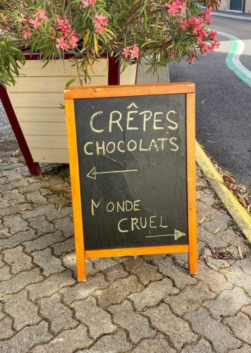 Photo of a chalkboard outside on a pavement saying "Crêpes chocolats" with arrow pointing left and "Monde cruel" with an arrow pointing right.