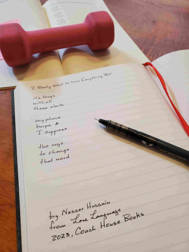 Handwritten transcription of the poem "I Really Want to Love Everything, But" from the poetry collection Love Language by Nasser Hussain - book is held open with a small pink hand weight
