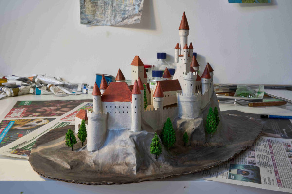 A model of a medieval castle made of cardboard, papier-mâché and small plastic trees, painted with acrylic paint. The castle has various towers, most are round with a pointy roof.