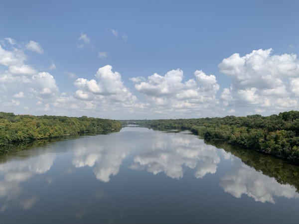 The calm waters of the Mississippi River reflect the blue sky and white puffy cumulus clouds. It has a green tree lined bank covering bluffs on each side.