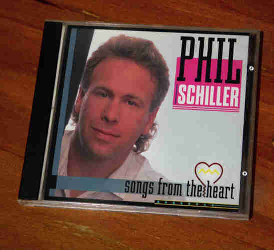 A photo of a CD jewel case of the album 'Songs from the heart' by Phil Schiller. Phil is peeking out seductively from behind a wall or something, with a lovely little mini mullet.