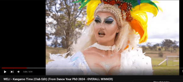 Still from linked video, shows a drag queen looking surprised in a flamboyant feathered costume. 