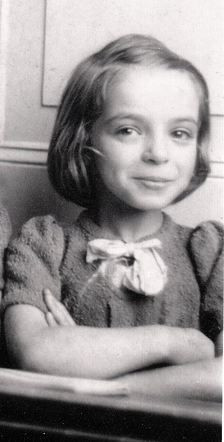 A vintage black and white photo of a young girl smiling, wearing a sweater with a bow on the collar, arms crossed in front of her.