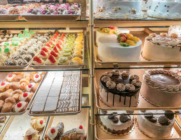 Dozens of cookies, many cakes and other colorful and fruit-topped pastries behind the glass of a well-stocked local bakery business. 