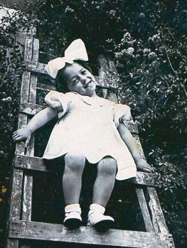 A young girl with a large bow in their hair sitting joyfully on a wooden ladder, in a vintage black-and-white photograph.