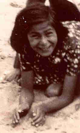 A girl on her knees on the sand. She leans on her elbows and scrapes the sand into her hands. She has long black hair. She is smiling broadly.