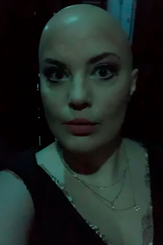 Gloomy, greenish image of a bald woman with heavy eye make-up and red lipstick looking into the camera. She is wearing several delicate gold necklaces. Selfie photo of Megan Marie Hart in costume and make-up as Chrysothemis, taken below the stage during a performance of Karsten Wiegand's staging of Elektra by Richard Strauss and Hugo von Hofmannsthal at Staatstheater Darmstadt, 2024.