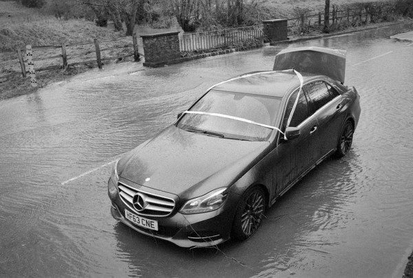 A stuck car is the main subject here, wrapped in Police tape and with its boot (or trunk?) lid up, it sits on a road still covered in an inch or so of water flooding from the brook, top centre. Black and white photo.